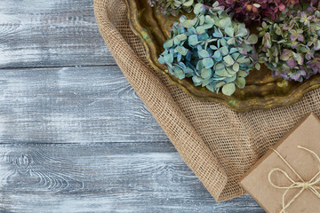 Vintage tray with dried blue flowers of hydrangeas, gift box wrapped in kraft paper on a burlap on a gray table. Flat style. Copy space for text.