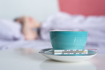 A cup of tea, thermometer with high body temperature and flu sick woman in bed background.