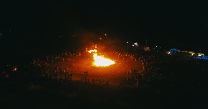 A large crowd of people celebrate near a giant bonfire. Burning hot sparks rise from large fire in the night sky. Belarusian Kupalle. Russian ethnic holiday Ivan Kupala in village. Aerial drone shot.