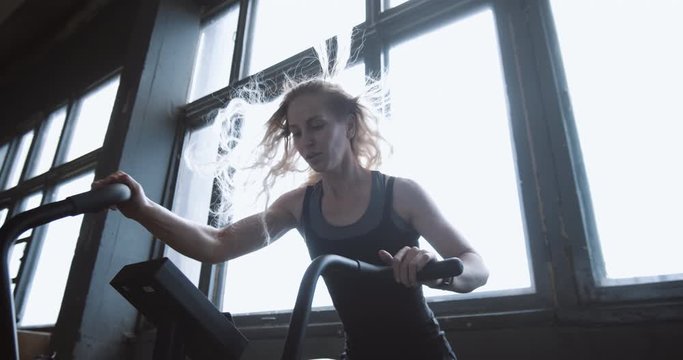 Young beautiful athletic blonde woman working out on stationary gym bike machine with wind in hair from fan slow motion.