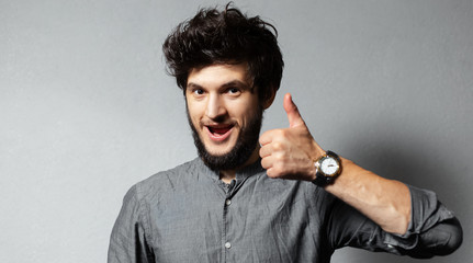 Portrait of young bearded guy with disheveled hair, show thumbs up on background of grey.