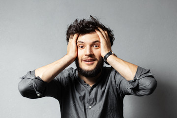 Portrait of young bearded guy with disheveled hair, amazed expression, holds hands on head.