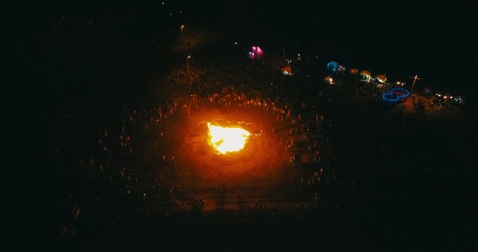 A large crowd of people celebrate near a giant bonfire. Burning hot sparks rise from large fire in the night sky. Belarusian Kupalle. Russian ethnic holiday Ivan Kupala in village. Aerial drone shot.
