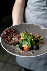 Waiter is holding flank steak with green salad, cherry tomatoes, grated nuts and black rice crisps with one hand