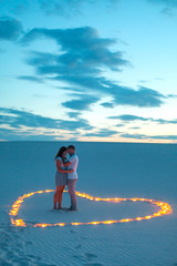 Couple in love romantic hugs in sand desert. Evening, romantic atmosphere, in sand burn candles in form of heart.