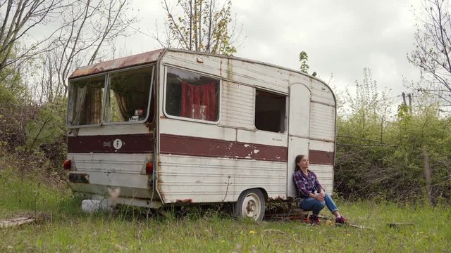 Young woman in casual wear meditatively sits near abandoned old caravan