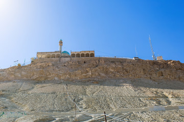 A mosque with a minaret and a blue dome on top of a rocky mountain in the desert against a blue...