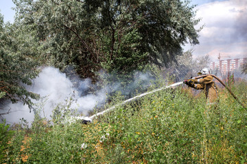 fireman in a protective suit with a hose pours water under burning pressure burning garbage in a natural landfill environment.