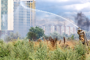 firefighter in a protective suit with a hose pours water over dry grass rescuing from a fire in the...