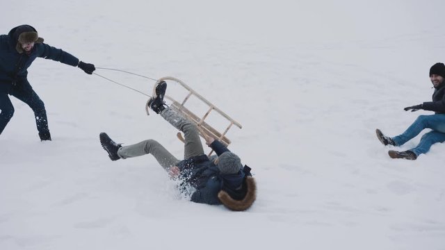 Happy friends playing on winter nature, pulling sled and turning over snow