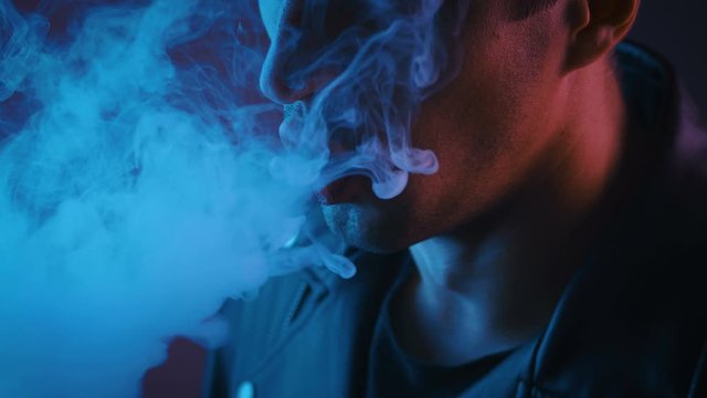 Man in leather black coat smoking vape or electronic cigarette on the blue and pink neon light background.