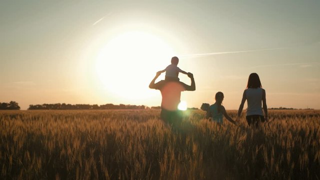 Happy family walking in wheat field on sunset in a warm summer evening. Little son sitting on her father shoulders, daughter holds mother hand and favorite toy. Concept of friendly family.