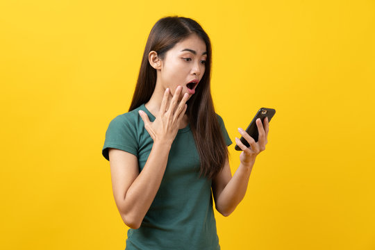 Shocked asian teenager in green tee shirt looking at mobile phone and having shock face on yellow background in studio.
