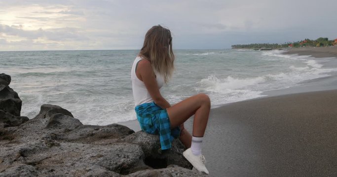 Young woman in modern outfit and checkered shirt sit on stone on the ocean black sand beach. Urban girl holiday. Summer vacation concept. Internet online freelance work break. Sport lifestyle.