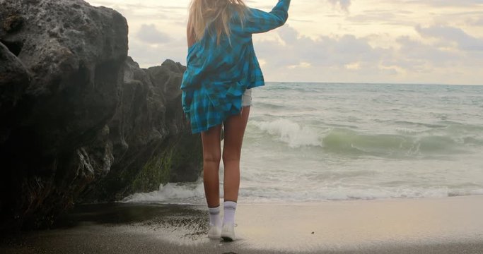 Young woman in modern outfit and checkered shirt with trendy backpack walk by a sand beach. Tourist urban girl holiday. Summer vacation concept. Internet online freelance work break. Sport lifestyle.