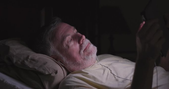 A mature man lying in bed engaged in the unhealthy habit of using an electronic tablet pc at bedtime which can negatively impact sleep patterns.