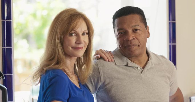 Happy Senior mixed race couple homeowners looking at camera. Older Caucasian and African American husband and wife standing in their kitchen smiling. Slow motion 4k handheld