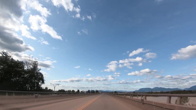 view from the windshield of a car moving along a highway and a bridge over a river, a huge sky with white clouds in the background