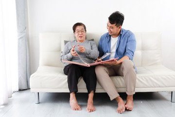 Asian senior woman mother and young man son in blue shirt looking photo album and talking happy smile face  in living room