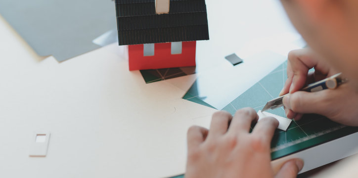 Close-up view of young male architect making small house model while cutting the cardboard