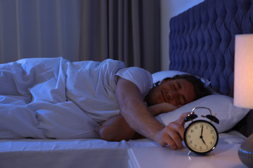 Sleepy young man turning off alarm clock on nightstand. Bedtime