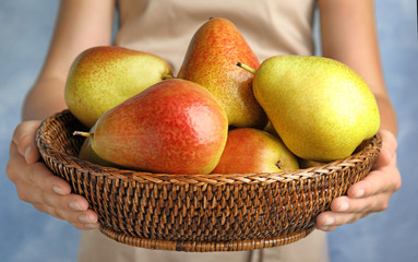 Woman holding wicker basket with ripe juicy pears on blue background, closeup