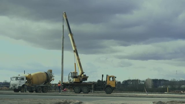 Workers install a pole with street light by automobile crane along the road. Construction machinery, concrete or cement mixer transport truck are on the work zone. Engineers set up lamppost on highway