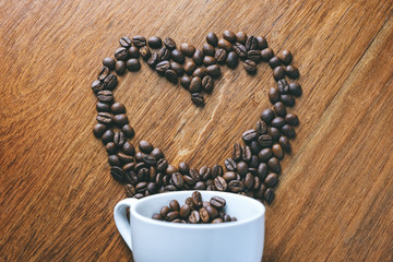 Top view image of coffee bean pouring out of a white mug to be a heart shape on wooden table