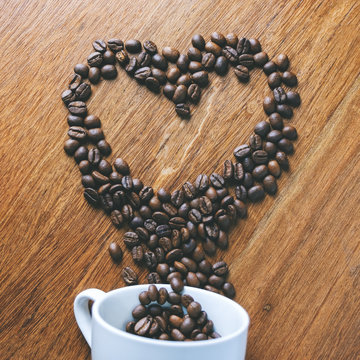 Top view image of coffee bean pouring out of a white mug to be a heart shape on wooden table