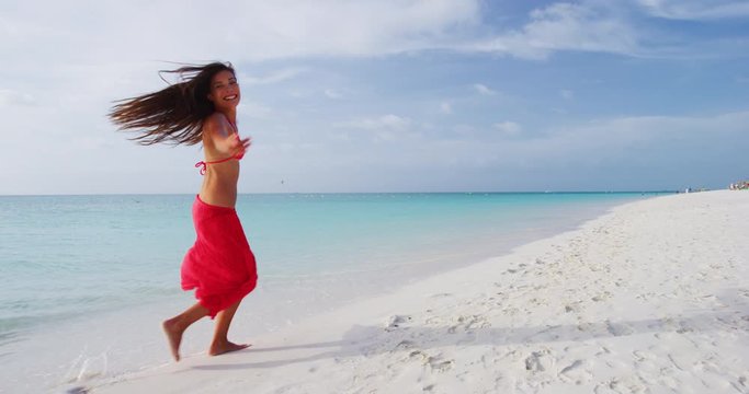 Beach Travel Joy and Happiness. Happy beach woman having fun running dancing on beach - weight loss or travel concept. Joyful girl wearing red bikini and sarong jumping of joy in beachwear. Slow mo