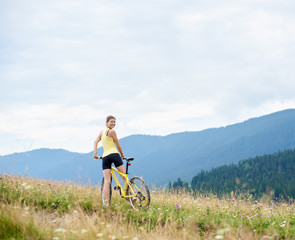 Back view of attractive happy woman cyclist riding on yellow mountain bicycle on a grassy hill, enjoying summer day in the mountains. Outdoor sport activity, lifestyle concept