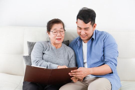 Asian senior woman mother and young man son in blue shirt looking photo album and talking happy smile face  in living room