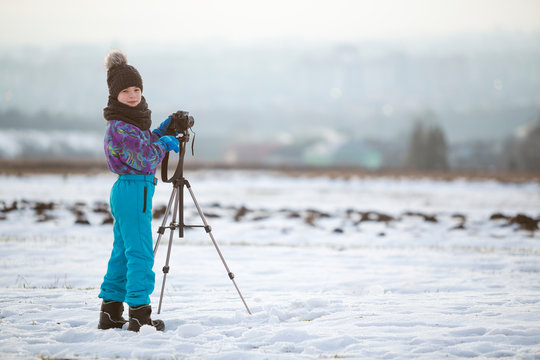 Child Boy Taking Pictures Outside In Winter With Photo Camera On A Tripod On Snow Covered Field.