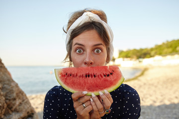 Young dark haired pretty woman in headband making fun over seaside view with slice of watermelon in her hands, looking at camera with wide eyes opened and raising eyebrows