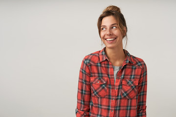 Studio shot of happy dark haired young female with bun hairstyle posing over white background, wearing casual clothes, looking aside with cheerful smile