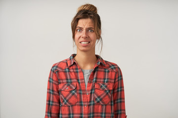 Studio photo of dark haired lovely woman standing over white background with confused face, wearing casual clothes, showing teeth and wrinkling forehead