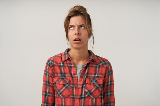 Puzzled young dark haired pretty female with casual hairstyle standing over white background, wearing checkered shirt and t-shirt, looking upwards and frowning