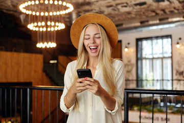Young positive long haired blonde female standing over restaurant interior in white shirt and brown hat, keeping smartphone in hands and looking at screen cheerfully