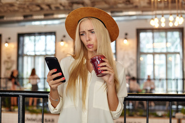 Indoor photo of beautiful confused woman with long hair posing over restaurant interior with cup of lemonade in hand, looking at her mobile phone and reading messages