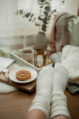 Toned photo. Autumn decor. Women's legs in knitted socks, a laptop, a mug of hot tea, a bun, candles. Cozy. Autumn.