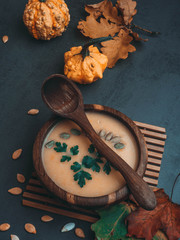wooden bowl of traditional pumpkin soup on gray table, decorated with seeds, parsley, colorful foliage, decorative pumpkin