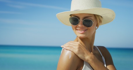 A happy young blond hair woman with a hat and sunglasses is applying a sunscreen or sun tanning...