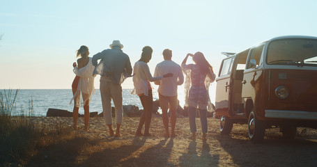 A group of young multi-ethnic friends just arrived with a minivan are having fun to start a vacation a beach with a sea on a sunset.