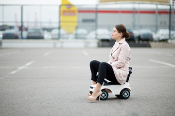 girl in a coat and trousers sits on a plastic car in a supermarket parking lot and looks in front...