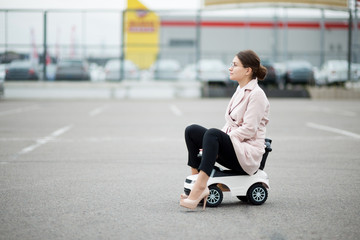 girl in a coat and trousers sits on a plastic car in a supermarket parking lot and looks in front of herself