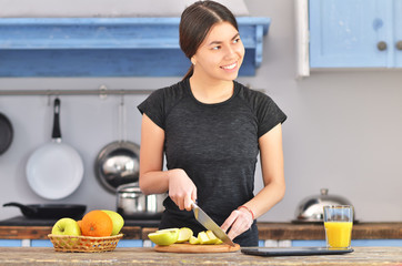 A young girl in a black t-shirt makes a fruit breakfast and cuts apple on a light wooden board in a kitchen.