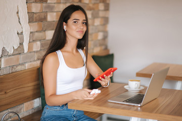 Attractive young girl use wirreless headphones and workin on her laptop. Woman in white t-shirt and denim. Student study in cafe
