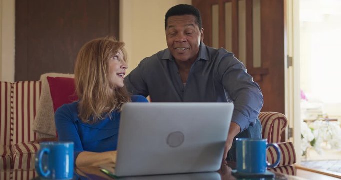 Senior African American And Caucasian Husband And Wife Use Laptop Computer Together In Living Room. Older Homeowner Married Couple Smiling While Booking Trip Online. Slow Motion 4k Handheld