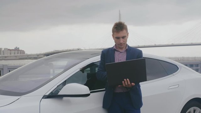 Concentrated businessman leans on electric car, holds laptop and works on it