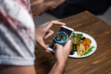 Men making photo (on his phone) of a delicious grilled chicken fillet and eggplant on a plate. Dark wooden table as a background
